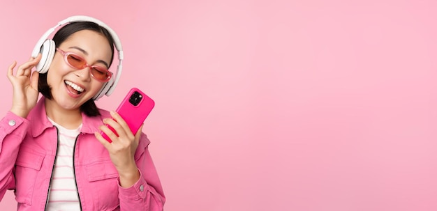 Stylish asian girl dancing with smartphone listening music in headphones on mobile phone app smiling and laughing posing against pink background