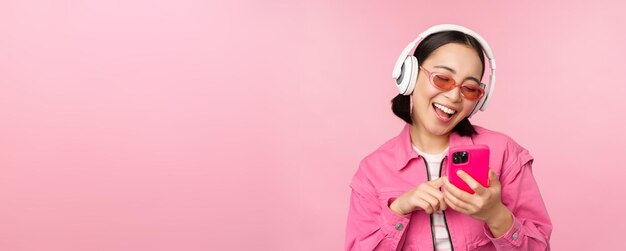 Stylish asian girl dancing with smartphone listening music in headphones on mobile phone app smiling and laughing posing against pink background