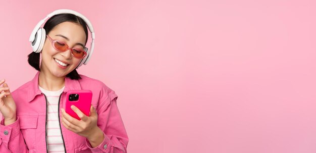 Stylish asian girl dancing with smartphone listening music in headphones on mobile phone app smiling and laughing posing against pink background