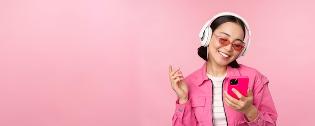 Stylish asian girl dancing with smartphone listening music in headphones on mobile phone app smiling