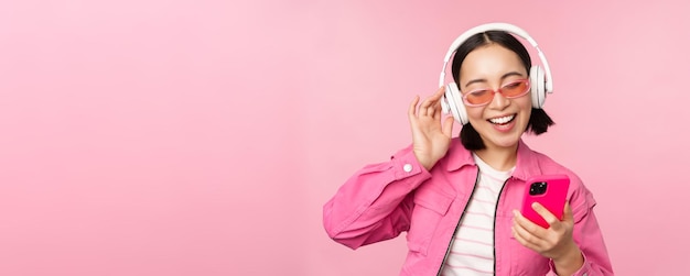 Stylish asian girl dancing with smartphone listening music in headphones on mobile phone app smiling