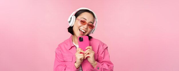 Stylish asian girl in headphones listening music and taking photos on mobile phone using smartphone standing over pink background