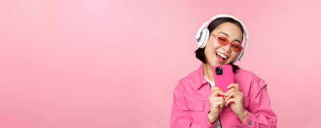 ピンクの背景の上に立っているスマートフォンを使用して音楽を聴いたり、携帯電話で写真を撮るヘッドフォンでスタイリッシュなアジアの女の子