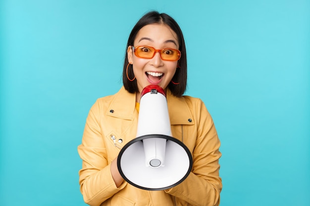 Бесплатное фото Стильная азиатская девушка делает объявление в мегафон, кричит по громкой связи и улыбается, приглашая людей набирать людей, стоящих на синем фоне