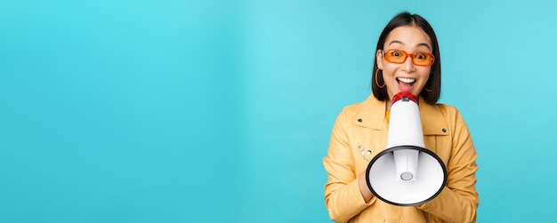 Stylish asian girl making announcement in megaphone shouting with speakerphone and smiling inviting