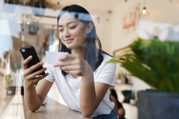Стильная азиатская девушка сидит в кафе у окна, оплачивая покупки в Интернете с помощью кредитной карты.