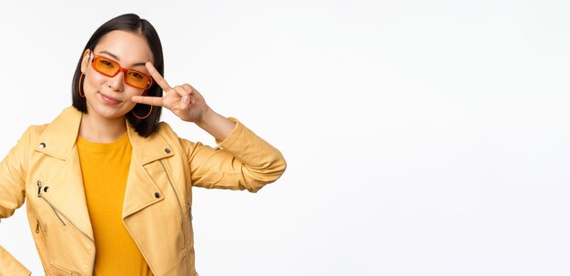 선글라스를 쓴 세련된 아시아 소녀는 시원하고 트렌디한 흰색 배경 위에 평화 vssign 제스처를 보여줍니다.