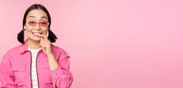 Stylish asian girl in sunglasses looks thoughtful or intrigued wants to try smth out stands over pink background pondering