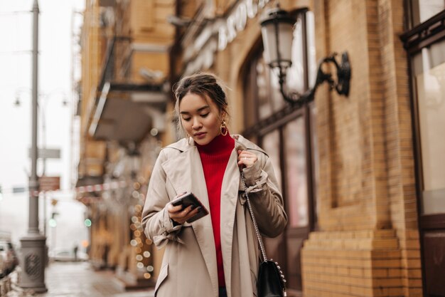 Стильная азиатка в бежевом пальто, красном топе и сумке через плечо гуляет по городу, держа в руках смартфон.