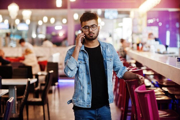 Stylish asian man wear on jeans jacket and glasses posed against bar in club and speaking on mobile phone