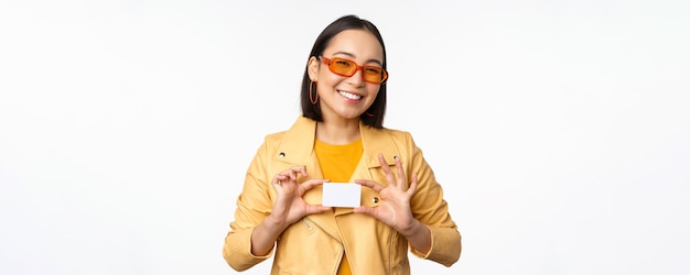 Stylish attractive asian girl in sunglasses showing credit card and smiling standing happy against white studio background