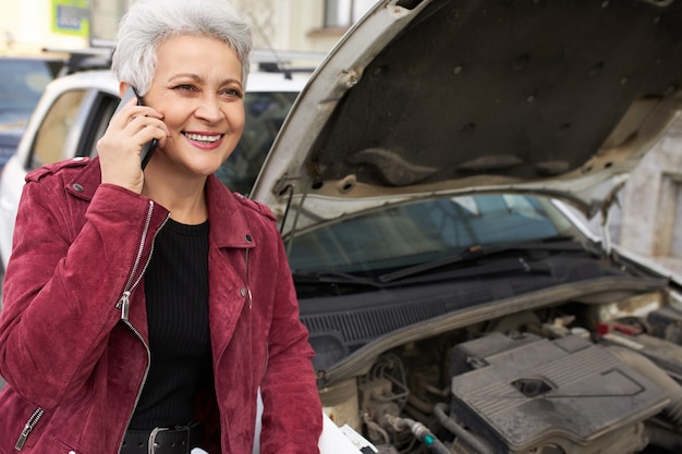 Бесплатное фото Стильная привлекательная седая зрелая женщина-водитель стоит возле своей разбитой белой машины с открытым капотом и разговаривает по телефону