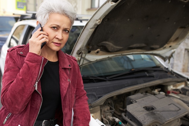 Stylish attractive gray haired mature female driver standing near her broken white car with open hood and talking on the phone