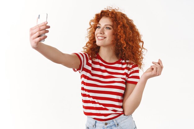スタイリッシュで魅力的な赤毛の縮れ毛の女の子ファッショナブルなストライキポーズ自撮りホールドスマートフォンを見せてソーシャルメディアのフォロワーに新しい夏の服を笑顔で広く立っている白い壁