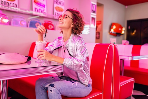 Стильная привлекательная улыбающаяся женщина в ретро винтажном кафе 50-х годов розового цвета сидит за столом