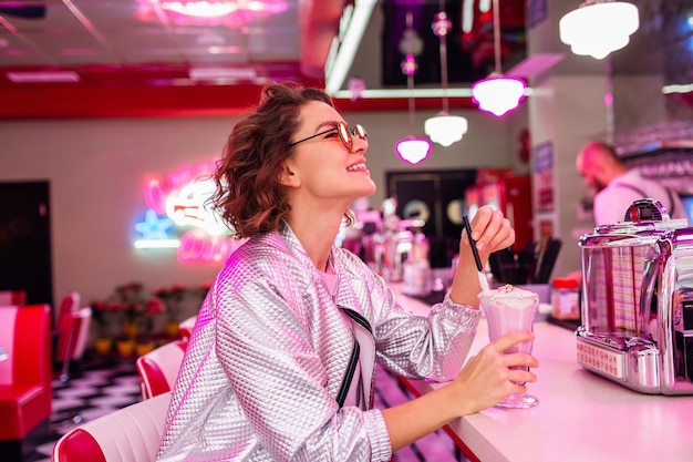 Stylish attractive smiling woman in retro vintage 50's cafe of pink color sitting at table