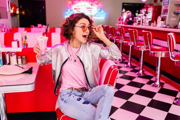 Stylish attractive smiling woman in retro vintage 50s cafe sitting at table drinking milk shake cocktail in hipster outfit having fun laughing in cheerful mood funny surprised face expression