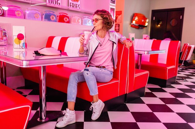 Stylish attractive smiling woman in retro vintage american 50s cafe sitting at table drinking milk shake cocktail in hipster stylish