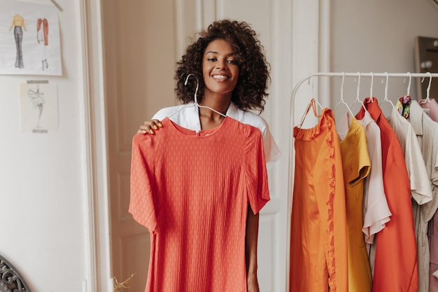 Stylish attractive woman in white blouse smiles sincerely and holds hanger with red dress in dressing room