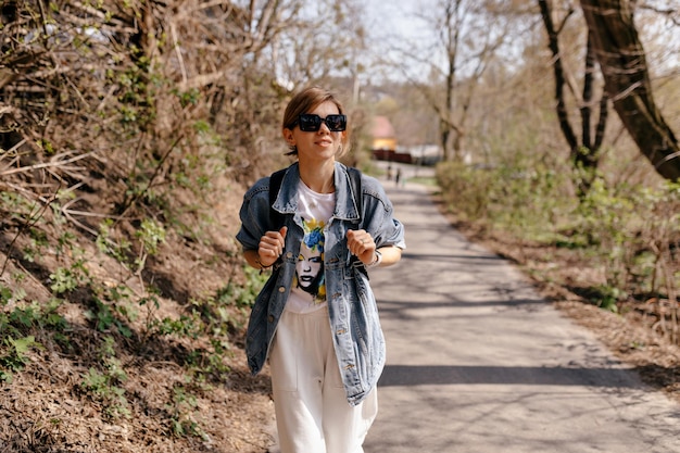 Free photo stylish attractive woman with collected hair wearing white pants and denim jacket is walking with backpack on forest trail in sunlight