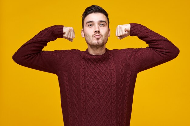 Stylish attractive young bearded guy in knitted cozy sweater demonstrating strength, raising hands, keeping fists clenched, feeling confident and proud of himself. Confidence and power concept