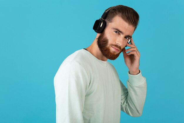 파란색 벽에 고립 된 무선 헤드폰 현대적인 스타일 자신감 분위기에서 음악을 듣고 세련 된 매력적인 젊은 수염 난된 남자