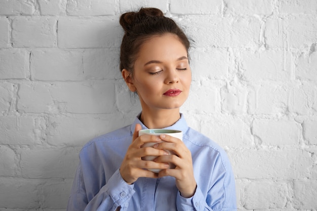 フォーマルな青いシャツを着て、ホットコーヒーを飲みながら目を閉じて、新鮮な香りを楽しんで、幸せそうに笑ってメイクアップスタイリッシュな魅力的な若いブルネットの女性サラリーマン