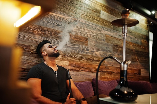 Stylish beard arabian man in glasses and black tshirt smoking hookah indoor bar Arab model having rest