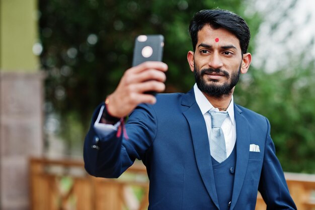 屋外でポーズをとって携帯電話で自分撮りを作る青いスーツの額の上のbindiを持つスタイリッシュなひげのインドのビジネスマン