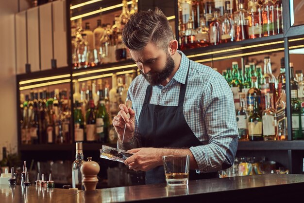 Stylish bearded bartender in a shirt and apron splits ice to make a cocktail at bar counter background.