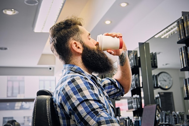 Un elegante hipster maschile vestito con una maglietta in pile, beve caffè nel salone di un parrucchiere.
