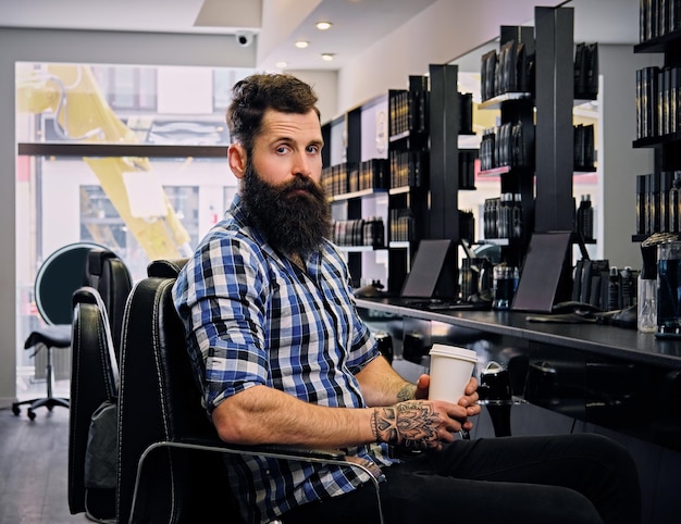A stylish bearded hipster male dressed in a fleece shirt, drinks coffee in a hairdresser's salon.