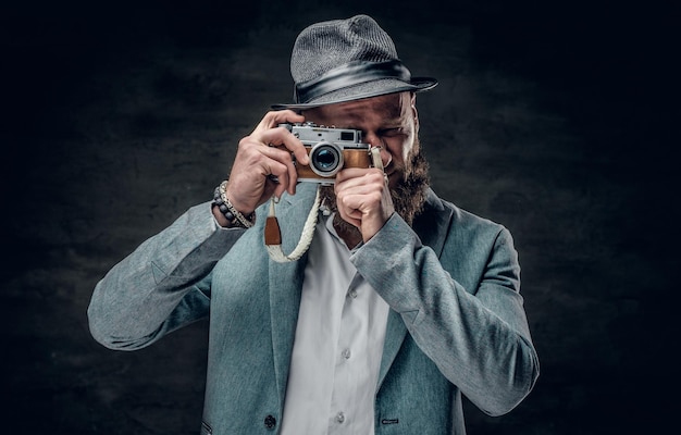 A stylish bearded hipster male dressed in a grey jacket and felt hat holds an SLR photo camera.