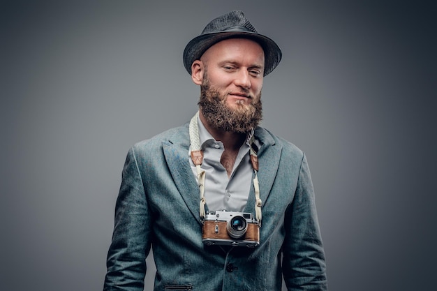 A stylish bearded hipster male dressed in a grey jacket and felt hat holds an SLR photo camera.