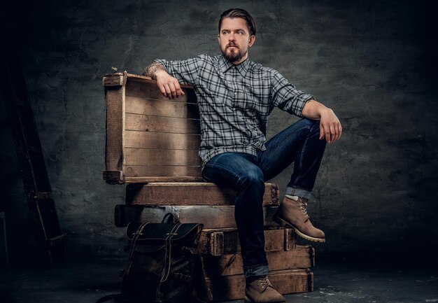 The stylish bearded hipster male sits on a wooden box in a studio over dark grey background.