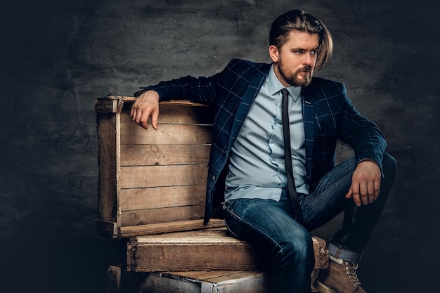 The stylish bearded hipster male sits on a wooden box in a studio over dark grey background.