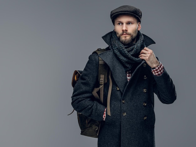 A stylish bearded hipster man dressed in a grey jacket holds a backpack.