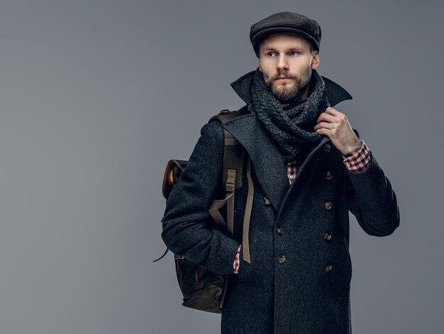 A stylish bearded hipster man dressed in a grey jacket holds a backpack.