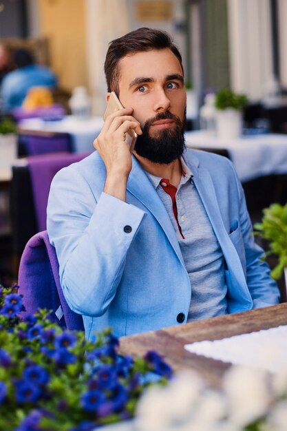 カフェのテーブルでスマートフォンを使用して青いジャケットを着たスタイリッシュなひげを生やした男性。