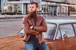 Free photo stylish bearded male dressed in brown leather jacket leaning on tuned retro car in the city parking.