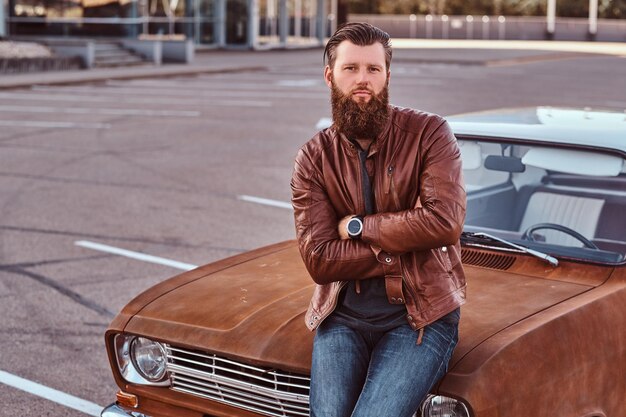 stylish bearded male dressed in brown leather jacket posing with crossed arms while leaning on tuned retro car in the city parking.