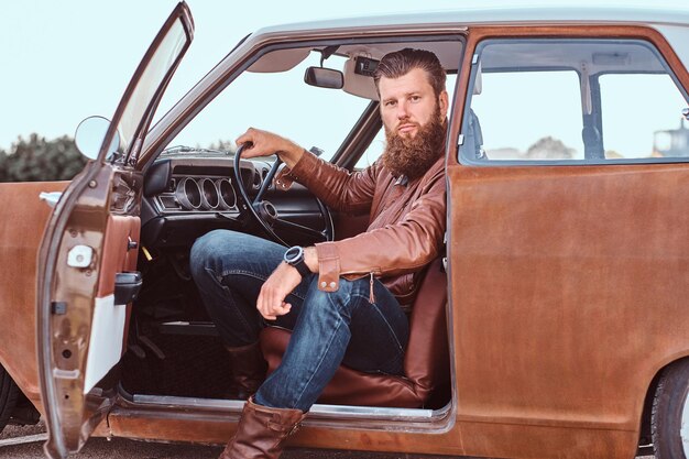 Stylish bearded male dressed in brown leather jacket sits behind the wheel of a tuned retro car with open door.
