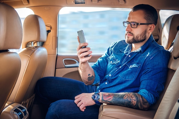 Free photo stylish bearded male in eyeglasses with tattoo on his arm, talking by smart phone in a car.