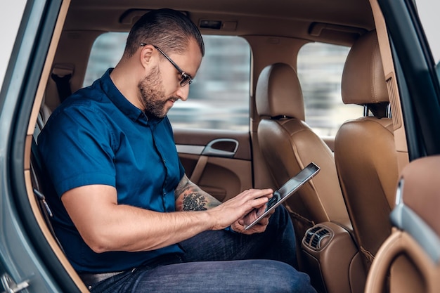 Stylish bearded male in eyeglasses with tattoo on his arm using portable tablet PC on a back seat of a car.