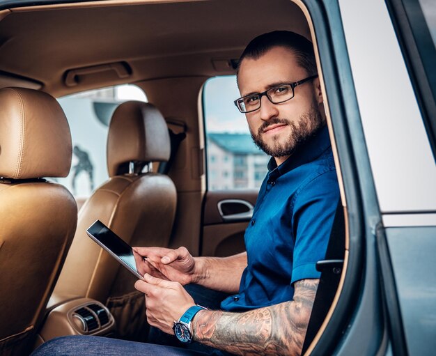 Stylish bearded male in eyeglasses with tattoo on his arm using portable tablet PC on a back seat of a car.