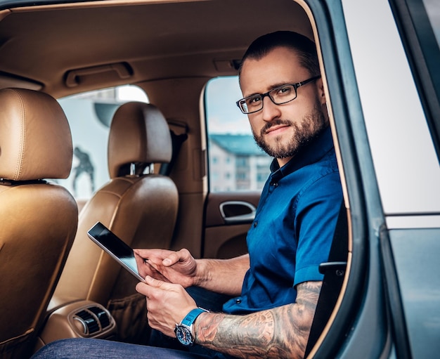 Стильный бородатый мужчина в очках с татуировкой на руке с помощью портативного планшетного ПК на заднем сиденье автомобиля.