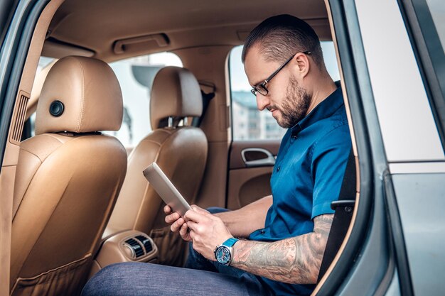 Stylish bearded male in eyeglasses with tattoo on his arm using portable tablet PC on a back seat of a car.