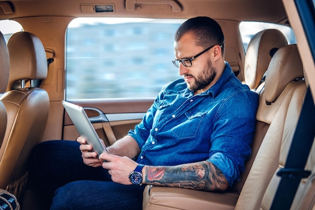 Free photo stylish bearded male in eyeglasses with tattoo on his arm using portable tablet pc on a back seat of a car.
