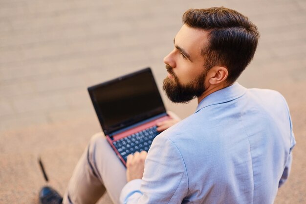 The stylish bearded male sits on a step and using laptop.