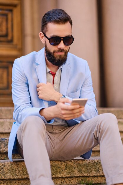 The stylish bearded male sits on a step and using smartphone.